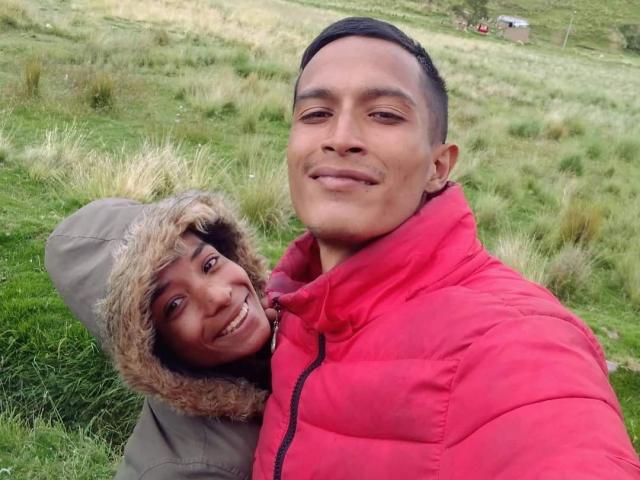 AlejoyMaria with short black hair, red puffy jacket, smiling, has his brown-skinned, curly-haired girlfriend in a green hooded jacket with fur trim, nuzzling his neck, in a grassy field.