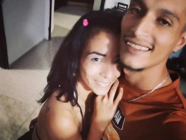 AlejoyMaria with dark hair and pink flower clip, smiling, next to a tan-skinned guy with a mustache, wearing a red shirt, in a selfie. Her hand on her cheek.