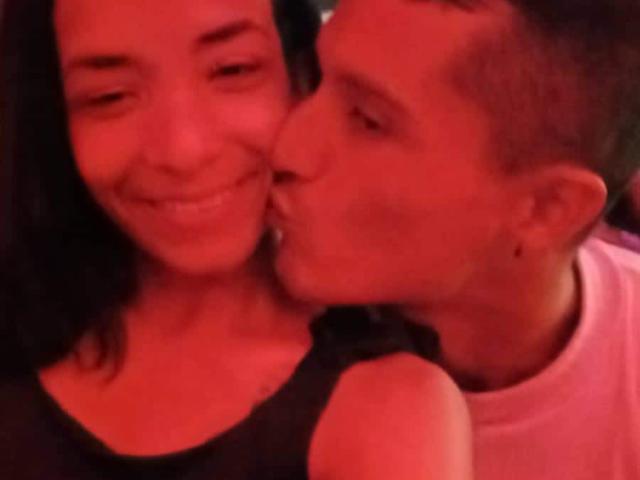 AlejoyMaria with dark hair and black top, smiling, has a man kissing her cheek. He's wearing a pink shirt, and the image is red-tinted. His face is near her tits.