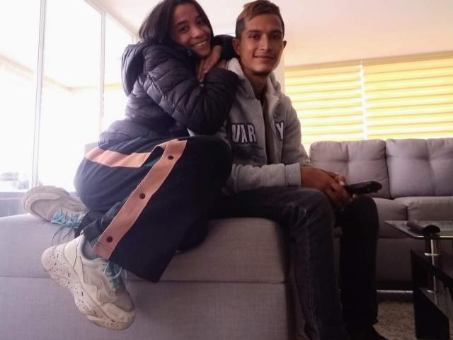 AlejoyMaria with dark hair, puffy black jacket, pink and black pants, white sneakers, sitting on sofa, smiling. AlejoyMaria with light brown hair, gray hoodie, holding game controller.