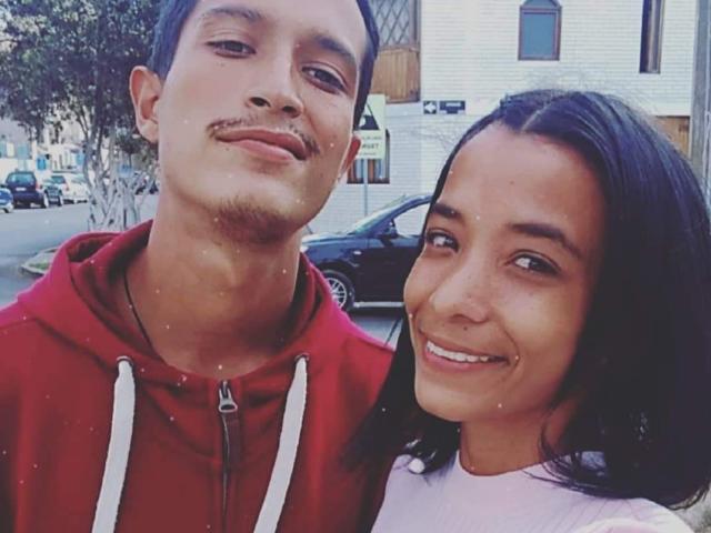 AlejoyMaria with short brown hair and a mustache, wearing a red hoodie, next to AlejoyMaria with long black hair, smiling, in a casual selfie. Both have cute faces.