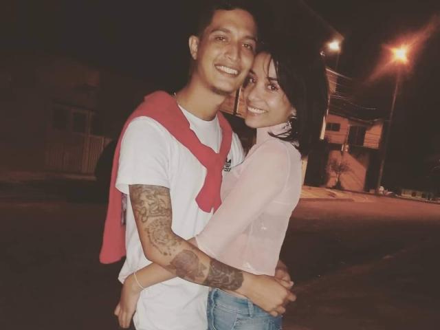 AlejoyMaria with tattoos on arm, white shirt, red scarf, smiling, hugging AlejoyMaria in pink top, blue jeans, dark hair, standing on street at night, arms around each other.
