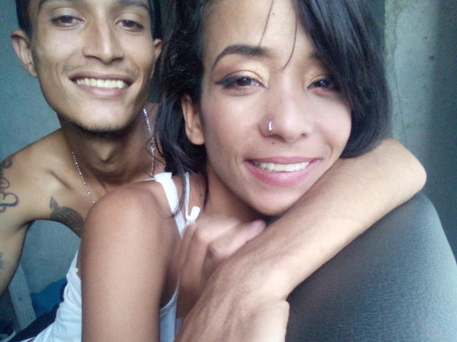 AlejoyMaria with nose ring, smiling, black hair, white tank top, arms around AlejoyMaria's shoulder, shirtless AlejoyMaria with tattoos, smiling, both have light brown skin, sitting together.