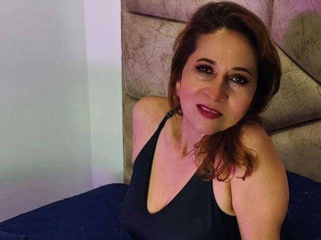MargarethRouss has red hair, dark eyes, and is wearing a black tank top, sitting with a slight smile, showing her tits and a hint of her ass.