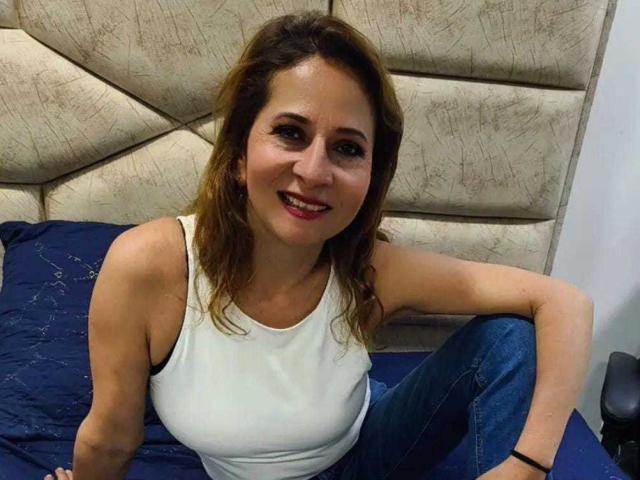 MargarethRouss, with brown hair and red lipstick, wearing a white tank top and blue jeans, sitting on a bed, smiling, showing her tits, with a black wristband.