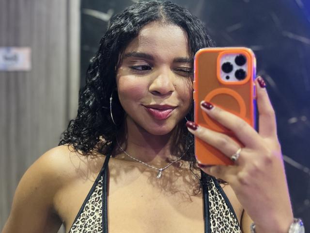 LisaMendez with wavy black hair, wearing a leopard print bikini top, holding an orange phone, winking, and smiling with red nails and a silver necklace.