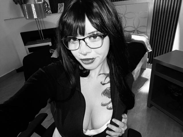 Selfie en blanco y negro de Natasha Hottson con gafas, cabello oscuro y un tatuaje en el pecho, mostrando sus senos en un top con escote pronunciado y la boca ligeramente abierta..