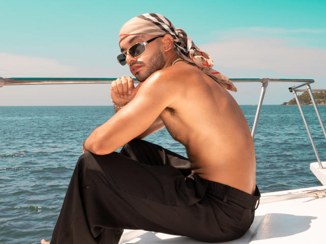 Valentino Bermont, zonder shirt, gekleed in zwarte broek, met zonnebril en een hoofddoek, zittend op een boot met een blauwe oceaan en een heldere lucht, kijkt om zich heen met een ontspannen, zelfverzekerde uitdrukking..