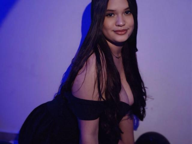 AlessLin, avec ses longs cheveux noirs, porte un haut noir à une épaule qui met en valeur sa poitrine généreuse, et elle est assise, souriante, avec une lumière bleue sur son visage. Ses fesses sont partiellement visibles..