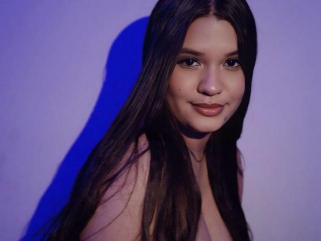 AlessLin with long dark brown hair, fair skin, and a small beauty mark near her nose, smiling softly with her tits barely visible, against a blue-lit background.