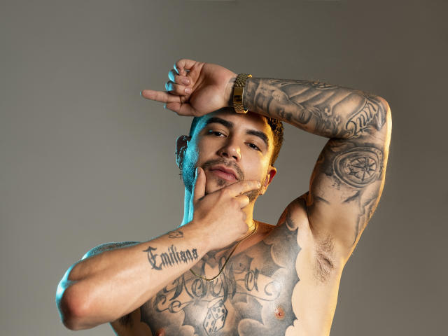 DanteDimarco, torse nu, avec des tatouages recouvrant sa poitrine et ses bras, une main sur le cou, un bras levé, éclairage bleu et orange, musclé, peau brune, cheveux courts et bouclés, expression confiante..