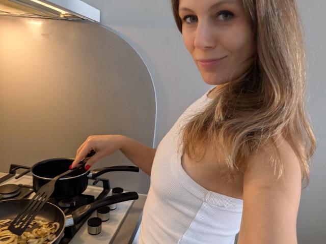 AuroraStar with blonde hair, white tank top, and red nails, cooking in a kitchen, stirring scrambled eggs in a pan with a black lid on a gas stove.