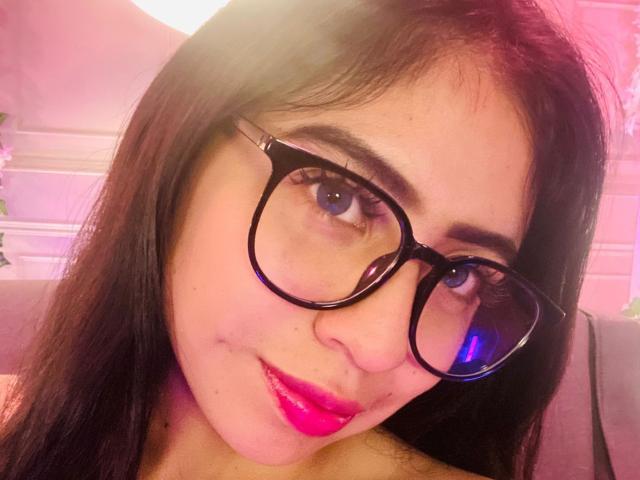 ZoeRedds, com óculos pretos, batom rosa e cabelo escuro de comprimento médio, olhando para a câmera com um leve sorriso. Ela tem olhos azuis e um tom de pele claro..