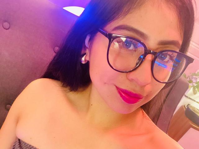 ZoeRedds with dark hair, big glasses, pink lipstick, and small earrings, showing her bare shoulders and a slight smile, looking into the camera.