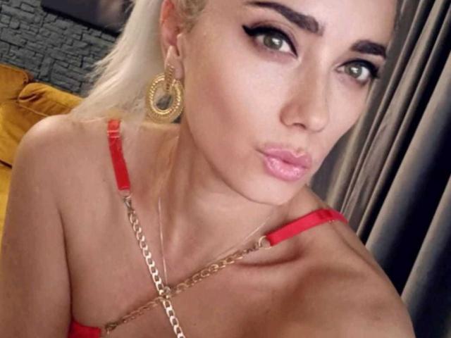 LaBelleAlice with pouty pink lips, green eyes, and thick eyebrows, wearing red lingerie with gold chain, and large hoop earrings, has blonde hair and a fair skin tone.