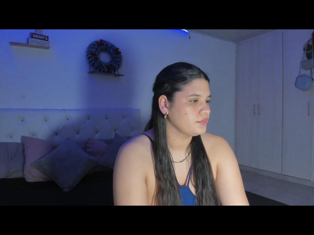 AlessLin with long black hair, wearing a blue tank top, looking to the side with a slight smile, showing small tits and a mole on her right cheek.