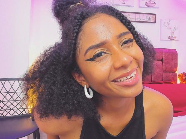 MeganWilliams with curly black hair, thick eyebrows, winged eyeliner, hoop earrings, smiling, showing teeth, wearing a black sleeveless top, in a brightly lit room.