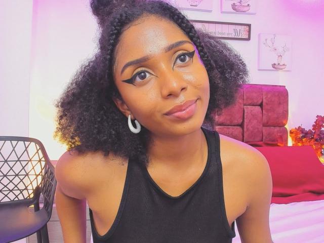 MeganWilliams has shiny brown skin, curly black hair in a bun, bold eyeliner, pink lipstick, hoop earrings, and wears a black sleeveless top, sitting on a red bed.