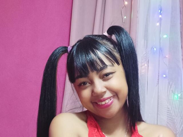 MaliciaQueen with black pigtails, pink bow, and red lace top, smiling at the camera, showing off her cute face and glossy pink lips, with colorful fairy lights.
