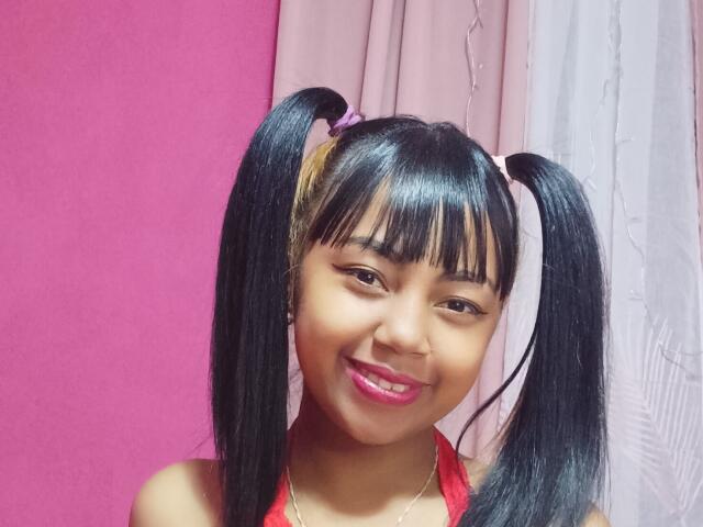 MaliciaQueen with black pigtails, pink lipstick, smiling, wearing a red top, brown eyes, straight black hair, pink and white hair ties, against a pink and white curtain.