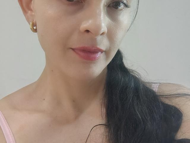 LatinaCulChaud has light skin, black hair in a ponytail, pink lipstick, and small gold earrings. She's smiling with a slight nose shadow, wearing a pink top.