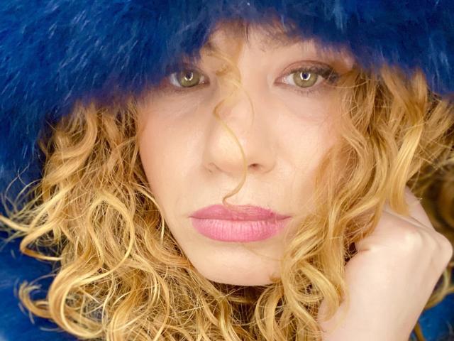 SweetJaqueline's face with curly blonde hair, blue furry hat, and pink lipstick, her green eyes staring intensely, hand touching her neck, looking serious and sexy as hell.