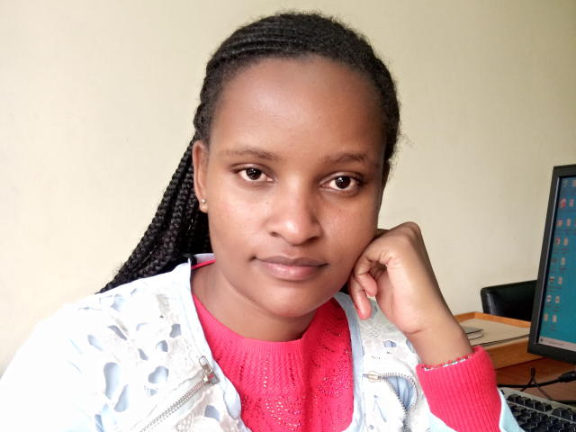 Emphi69, a Black woman with medium brown skin and small, round brown eyes, looks directly at the camera. She has long, tightly braided black hair. She wears a white, patterned, zip-up jacket over a bright pink, textured knit shirt. Her right hand rests on her cheek. In the background, a computer with a blue screen is on a desk to the right. The image is a close-up, and Emphi69's expression is neutral. The lighting is even, highlighting her natural skin tone.