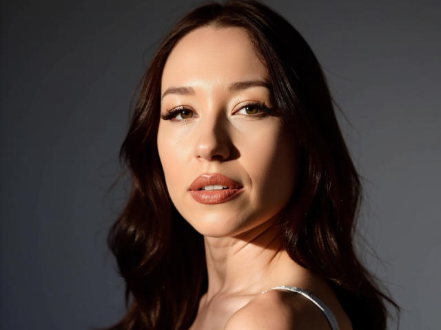 MartinaFlirt has brown wavy hair, light skin, and full, glossy pink lips. Her brown eyes have long, thick eyelashes. She's wearing a white strap, looking slightly to the side.