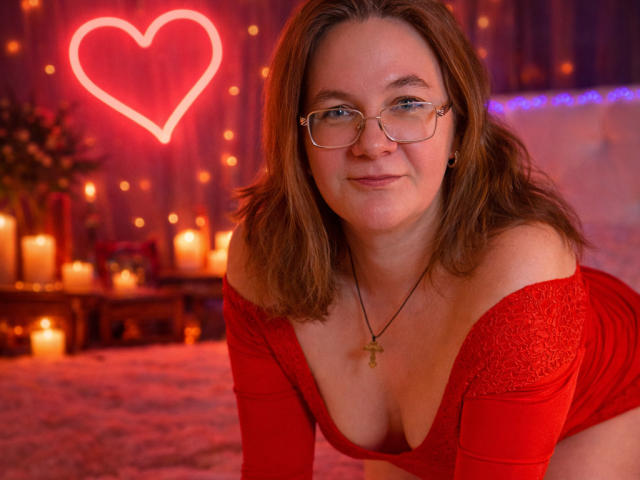 HolyMilfa with glasses, red off-shoulder top, showing tits, brown hair, cross necklace, smiling, candles, heart neon in the background.