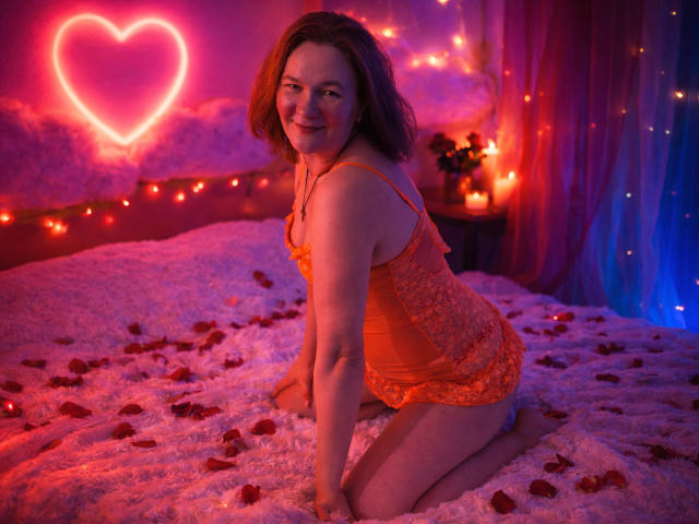 HolyMilfa, in an orange lace babydoll, kneels on a rose-petalled bed, smiling, with pink neon heart in the background, and soft, warm, red-blue lighting. Her tits are visible.