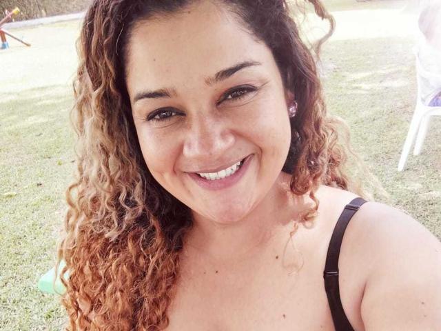Close-up of NataliaWendys with curly, light brown hair that fades into blonde at the tips. She has fair skin, brown eyes, and a wide smile showing white teeth. She wears a black strap over her shoulder. The image is bright with natural light highlighting her face. Her eyebrows are thick, and she has small, pink earrings. The background is blurred, focusing on NataliaWendys' cheerful expression.