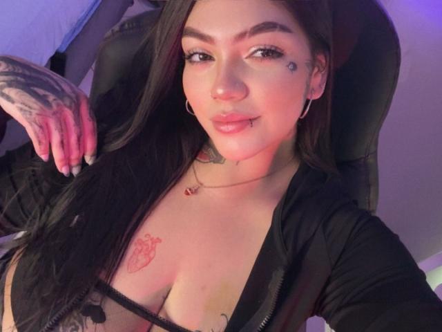 TessaMagic with long black hair, tattoos on her hand and chest, hoop earrings, and a black top, showing her tits in a selfie, pink lipstick, and a tongue piercing.
