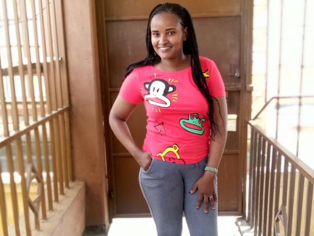 Emphi6 stands in a corridor with metal railings. She has dark skin and long braided hair. She wears a bright pink t-shirt with a cartoon monkey face and colorful graphics, and gray pants. She smiles, with her right hand in her pocket and a green bracelet on her left wrist. The corridor has wooden doors and light entering from the right.