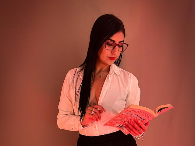 PaullieEvans with long black hair, red glasses, and red lipstick, wearing a white button-down shirt exposing her cleavage, black skirt, reading a book, looking down, red-tinted lighting.