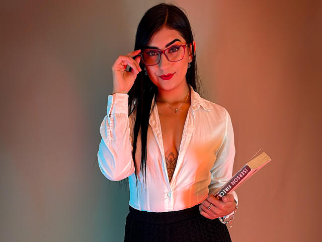 PaullieEvans with long black hair, red lipstick, and glasses, wearing a white button-up shirt, black skirt, holding a book, and showing a tattoo under the shirt.