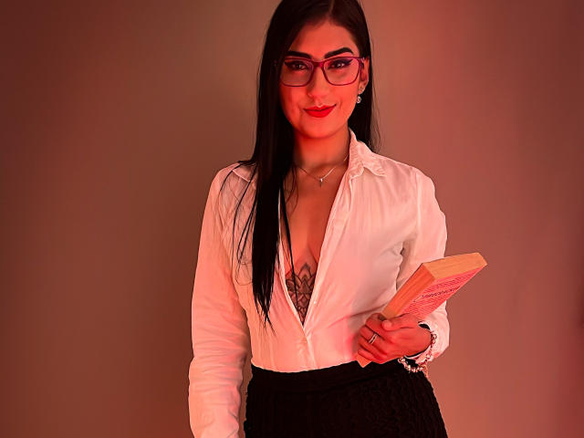 PaullieEvans with long black hair, red glasses, white unbuttoned shirt showing tits, black skirt, holding book, red lipstick, necklace, and bracelet, standing against warm light.