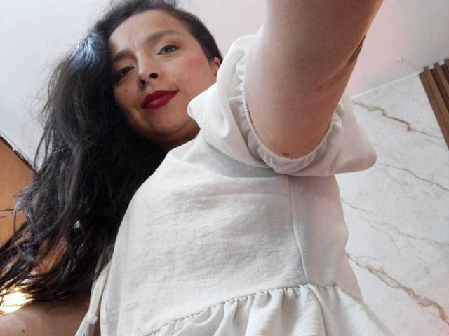 StangelarRivera with long black hair, red lipstick, and white blouse, taking a selfie from a low angle, showing her raised arm and smiling face. Boobs partially visible.