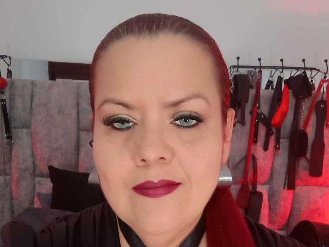 VanesaMoka with slicked-back brown hair, green eyeshadow, and red lipstick, wearing a black leather choker, standing in front of BDSM gear on a grey upholstered headboard.