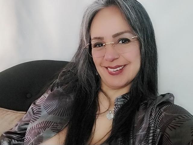 LailaCastillo with long black hair, glasses, smiling, wearing a patterned shirt, necklace with a moon charm, showing her tits slightly, sitting on a black cushioned chair.