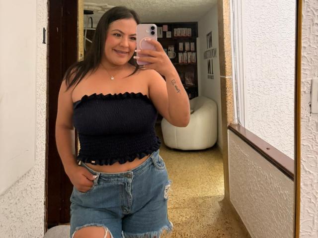 ScarlettOxford with dark hair, smiling, wearing a black strapless top, showing her tits, and ripped blue jeans, taking a mirror selfie with a pink phone.