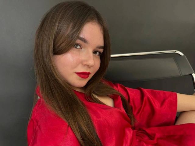 SofyX-hot has straight, medium-brown hair, parted to the side, and looks over her shoulder with a subtle smile. She wears a shiny red silk robe with a relaxed, confident posture. Her bright red lipstick contrasts with her fair skin. The image has a modern, slightly minimalist style with a focus on SofyX-hot's face and hair. The background is a dark, solid color, and the black chair she sits on has a sleek, modern design. The composition is centered on SofyX-hot's face, highlighting her relaxed expression and the texture of her hair and robe.