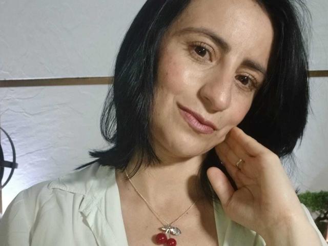BrendaCantorel with black hair, brown eyes, and a subtle smile, wearing a white shirt, red cherry necklace, and gold ring, touching her cheek.