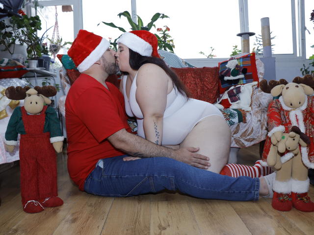 SarahMat with big ass, white tits, and a pussy in a white one-piece, kissing a bearded dude in a red shirt and jeans, both wearing Santa hats, on a wooden floor.