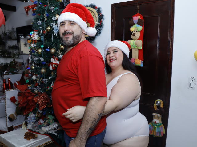 SarahMat, vêtue d'une chemise rouge et d'un bonnet de Père Noël, et portant des tatouages, se tient à côté d'une SarahMat plus corpulente, en justaucorps blanc et avec un bonnet de Père Noël, qui lui pose la main sur les fesses. Sapin de Noël..