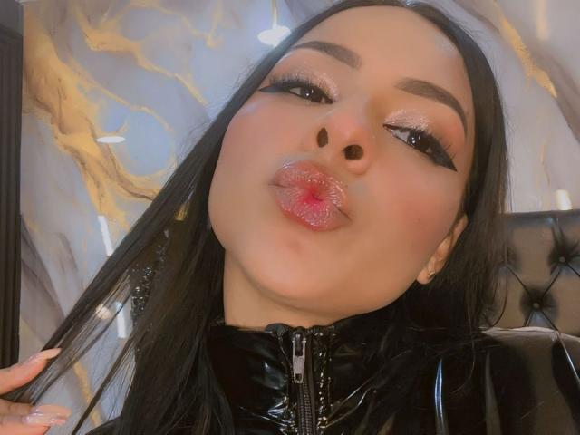 LindaFrancoo has glossy pink lips puckered, black eyeliner wings, and long black hair. She wears a black latex top with a zipper. Her skin is light.