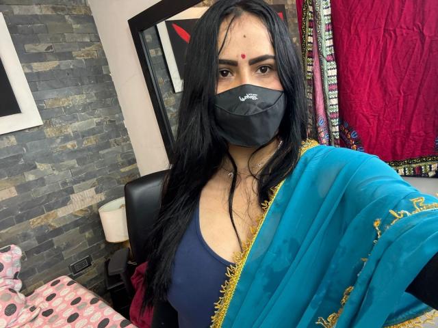 Dalia Dubey, con lunghi capelli neri, indossa un sari blu e dorato, una mascherina nera, un bindi rosso e una maglietta blu scuro, mostrando il seno e guardando la telecamera..