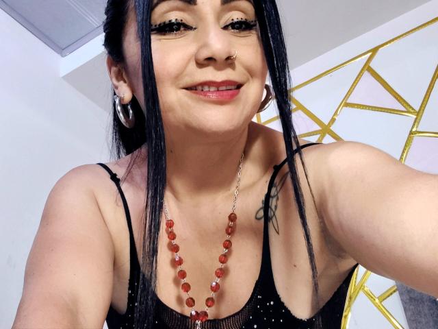 Zaria69, com cabelo preto, piercing no nariz e brincos de argola, sorrindo para a câmera, vestindo uma blusa de malha preta com um colar de contas vermelhas, mostrando uma tatuagem no braço direito..