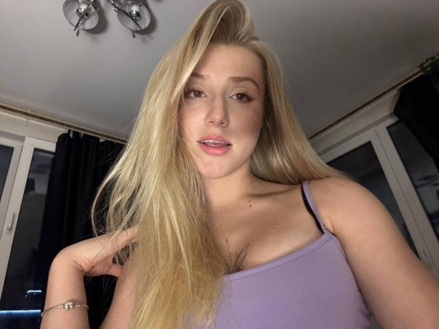 Blonde ArianaMur with long hair, pink lips, and light skin, wearing a purple tank top, showing her tits, looking at the camera with a slight smile.