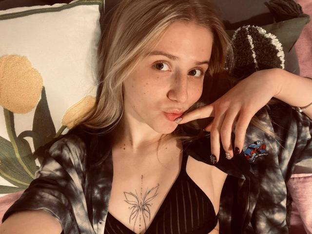ArianaMur with blonde hair, pouty lips, and a butterfly tattoo on her chest, wearing a black tie-top and a tie-dye shirt, looking at the camera.