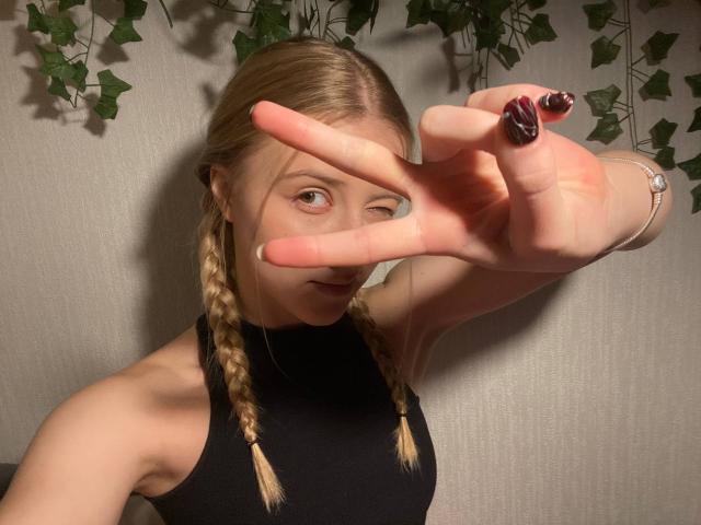 ArianaMur with blonde braids in black sleeveless top, flashing a peace sign with dark nail polish, showing her ass and a ring on her finger, in a dim room.